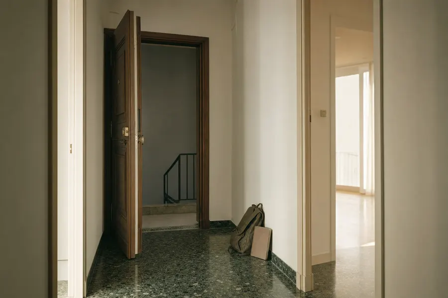 Recibidor vacío de un piso luminoso con la puerta abierta al rellano, una carpeta de documentos y una mochila apoyadas en el suelo