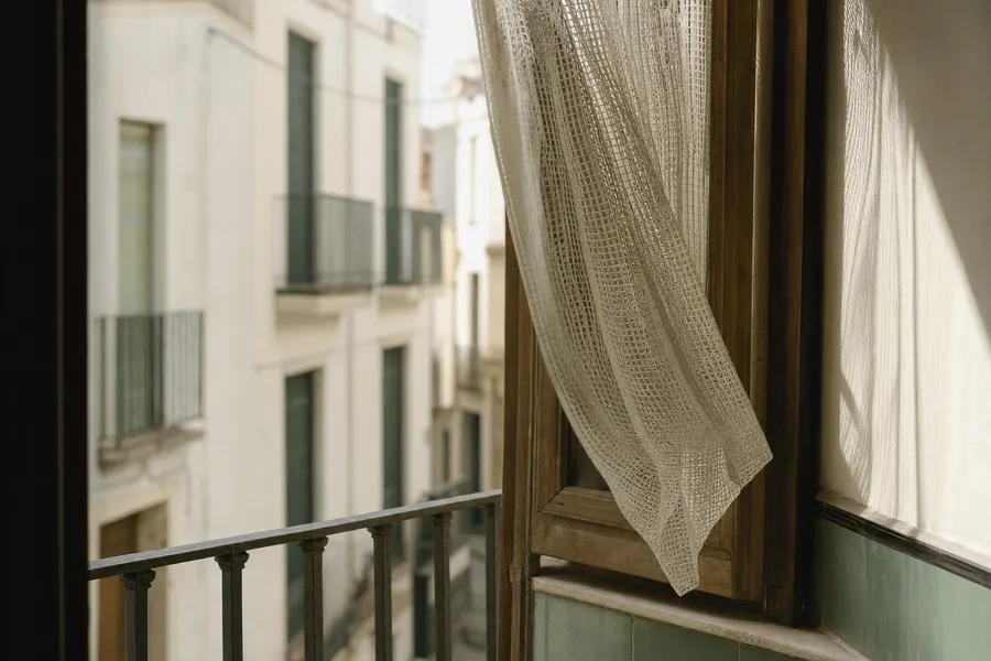 Visillo apartado en la ventana de un piso, con la calle desenfocada al otro lado